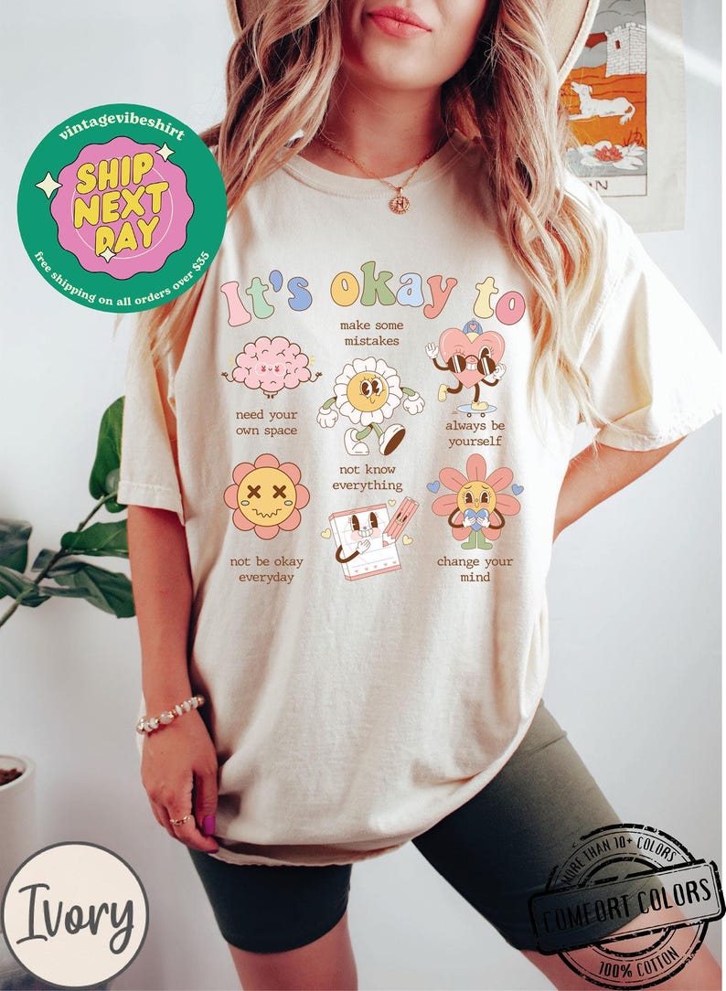 May include: A cream-colored t-shirt with the text "It's okay to" and a series of colorful illustrations of cartoon characters with positive messages. The illustrations include a brain, a flower, a heart, a flower with a sad face, a calendar, and a flower with a heart. The text below each illustration includes phrases like "make some mistakes", "need your own space", "not know everything", "always be yourself", "not be okay everyday", and "change your mind".