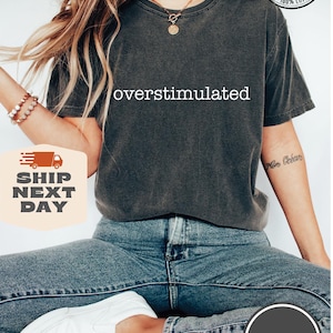 May include: Dark grey Comfort Colors t-shirt with the word "overstimulated" in white. The shirt is 100% cotton. The model is wearing blue jeans and white shoes. The image also includes the text "More than 10 colors" and "Pepper".