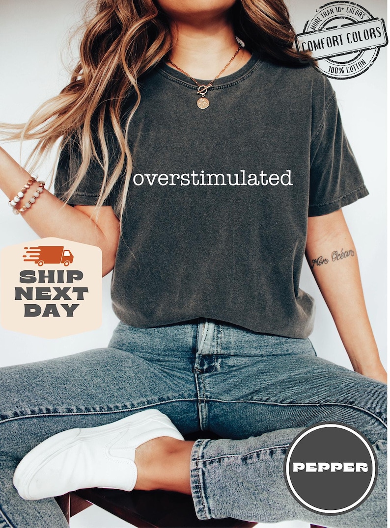 May include: Dark grey Comfort Colors t-shirt with the word "overstimulated" in white. The shirt is 100% cotton. The model is wearing blue jeans and white shoes. The image also includes the text "More than 10 colors" and "Pepper".
