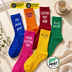 May include: A collection of six ribbed crew socks in blue, orange, magenta, dark red, yellow, and green. Each sock features white or black text including phrases like I'm just a gurl, MAYBE BABY, CUSTOM TEXT HERE, YOUR CUSTOM TEXT, BUY ME COFFEE, and it's a mood.