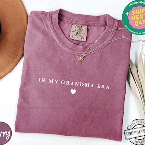 May include: A mauve colored t-shirt with the text "IN MY GRANDMA ERA" and a small white heart. The shirt is made of a soft, comfortable fabric.