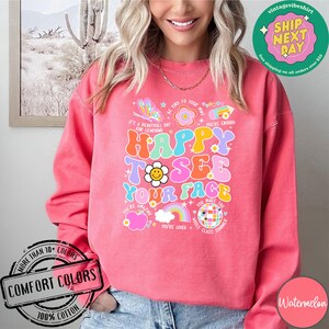 May include: Pink sweatshirt with a colorful graphic design that says "Happy to see your face". The graphic includes a rainbow, a smiley face, a disco ball, and other colorful elements. The sweatshirt is made of 100% cotton.