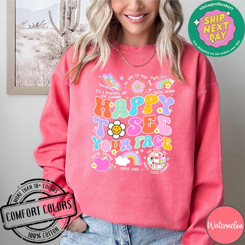 May include: Pink sweatshirt with a colorful graphic design that says "Happy to see your face". The graphic includes a rainbow, a smiley face, a disco ball, and other colorful elements. The sweatshirt is made of 100% cotton.