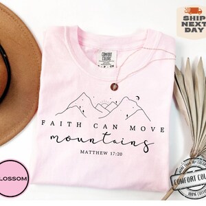 May include: Pink t-shirt with a black line drawing of mountains, a sun, and a moon. The text "FAITH CAN MOVE mountains MATTHEW 17:20" is printed on the shirt.