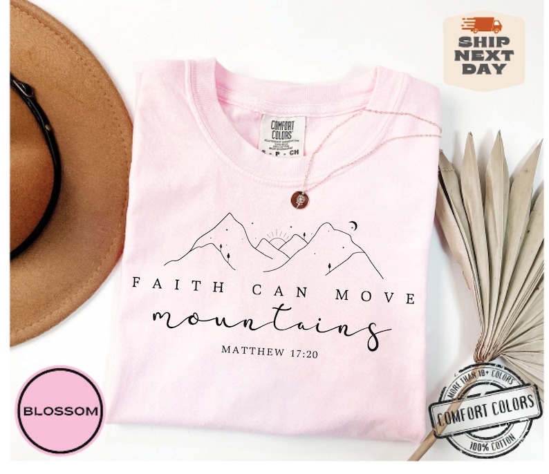 May include: Pink t-shirt with a black line drawing of mountains, a sun, and a moon. The text "FAITH CAN MOVE mountains MATTHEW 17:20" is printed on the shirt.