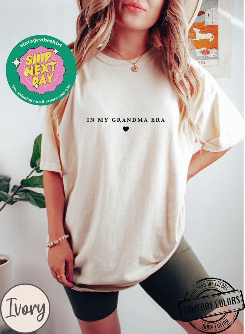 May include: A white t-shirt with the text "IN MY GRANDMA ERA" and a small black heart. The shirt is being worn by a person with long blonde hair.