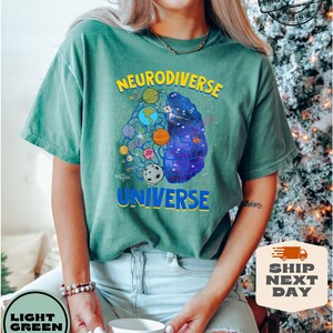 Op de afbeelding: Een lichtgroen T-shirt met een afbeelding van een brein met planeten en sterren erin. De tekst "Neurodiverse Universe" staat op het T-shirt gedrukt.