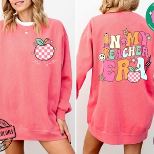 K&ouml;nnte beinhalten: Rosafarbener Pullover mit einem Grafikdruck eines Apfels mit kariertem Muster und dem Text "Mrs. Smith" auf der Vorderseite. Die R&uuml;ckseite des Pullovers zeigt eine Grafik mit dem Text "In My Teacher Era" in bunten Buchstaben mit einem karierten Apfel und anderen grafischen Elementen.