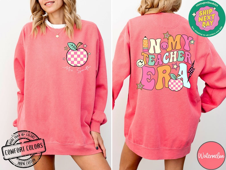 K&ouml;nnte beinhalten: Rosafarbener Pullover mit einem Grafikdruck eines Apfels mit kariertem Muster und dem Text "Mrs. Smith" auf der Vorderseite. Die R&uuml;ckseite des Pullovers zeigt eine Grafik mit dem Text "In My Teacher Era" in bunten Buchstaben mit einem karierten Apfel und anderen grafischen Elementen.