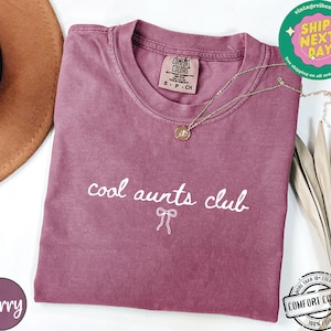 May include: A mauve colored t-shirt with the text "cool aunts club" in white lettering and a small white bow underneath. The shirt is made of a soft, comfortable fabric.