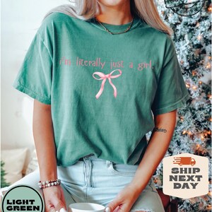 Puede incluir: Una camiseta verde claro con el texto "I'm literally just a girl." y un lazo rosa. La camiseta la lleva puesta una persona con un collar de oro y una taza blanca en la mano.