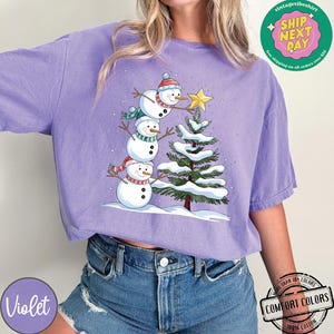 以下が含まれることがあります： 3人の雪だるまが積み重なっているデザインの紫色のTシャツ。一番上の雪だるまが緑色のクリスマスツリーの上に星を置いている。