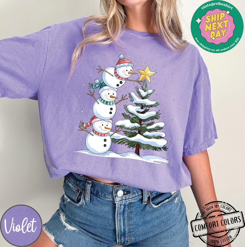 以下が含まれることがあります： 3人の雪だるまが積み重なっているデザインの紫色のTシャツ。一番上の雪だるまが緑色のクリスマスツリーの上に星を置いている。