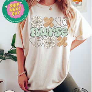 May include: A light beige t-shirt with a green "nurse" graphic. The graphic is surrounded by white daisies, red hearts, and brown bandages.