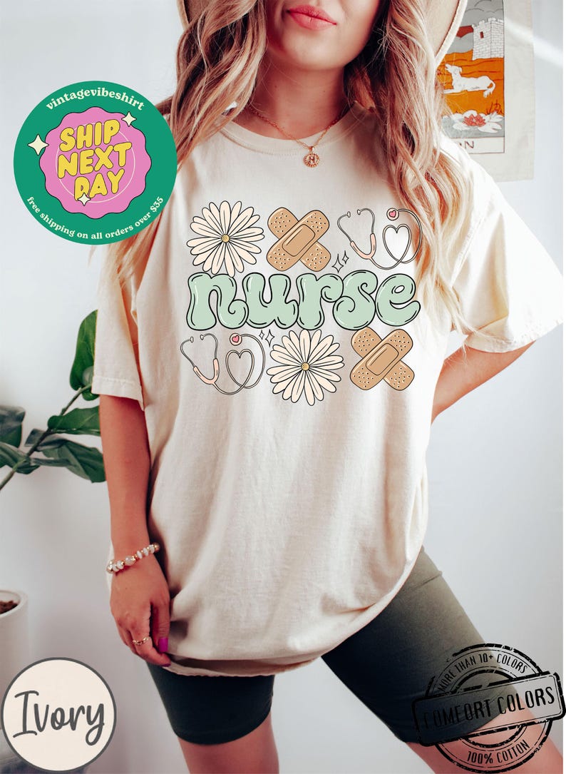 May include: A light beige t-shirt with a green "nurse" graphic. The graphic is surrounded by white daisies, red hearts, and brown bandages.
