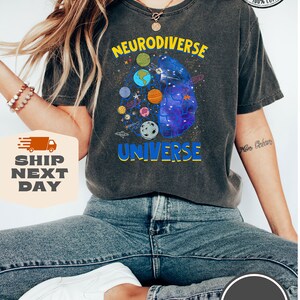 Op de afbeelding: Een donkergrijs T-shirt met de tekst "Neurodiverse Universe" in gele en blauwe letters. Het ontwerp toont een brein met een sterrenstelsel erin en planeten eromheen.