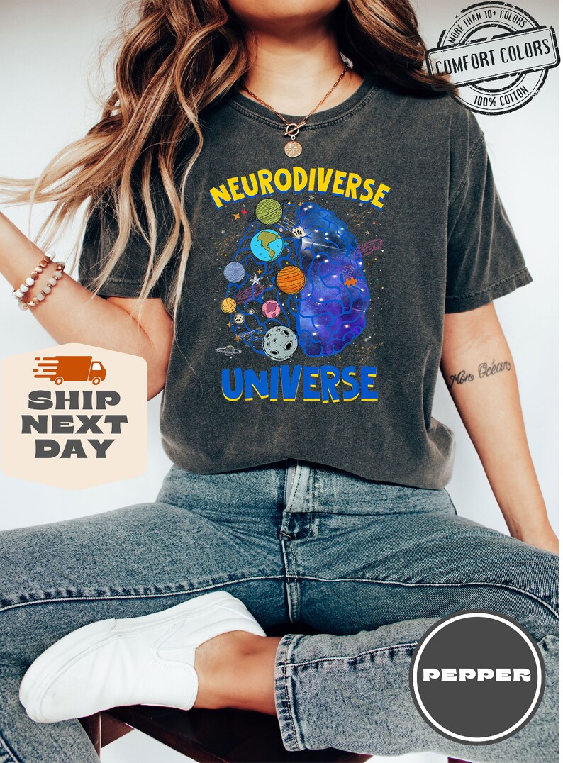 Op de afbeelding: Een donkergrijs T-shirt met de tekst "Neurodiverse Universe" in gele en blauwe letters. Het ontwerp toont een brein met een sterrenstelsel erin en planeten eromheen.
