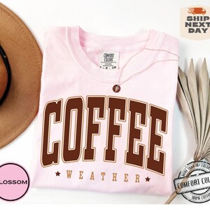 May include: Pink t-shirt with brown text that reads "COFFEE WEATHER" with a star on either side of the word "WEATHER".