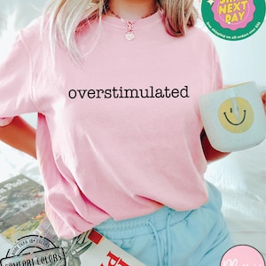 May include: A light pink Comfort Colors t-shirt with the word "overstimulated" printed in black. The image also shows a white mug with a yellow smiley face, and a book titled "How to Plan".