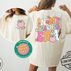 K&ouml;nnte beinhalten: Ein cremefarbenes T-Shirt mit einem grafischen Design aus einem bunten Apfel, einem Bleistift und dem Text "In My Teacher Era". Das Shirt ist in Elfenbein erh&auml;ltlich.