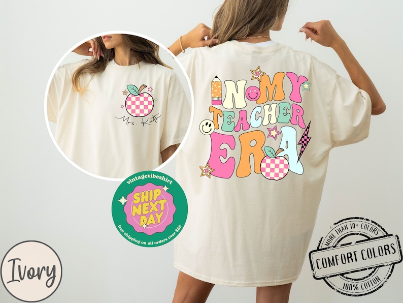 K&ouml;nnte beinhalten: Ein cremefarbenes T-Shirt mit einem grafischen Design aus einem bunten Apfel, einem Bleistift und dem Text "In My Teacher Era". Das Shirt ist in Elfenbein erh&auml;ltlich.