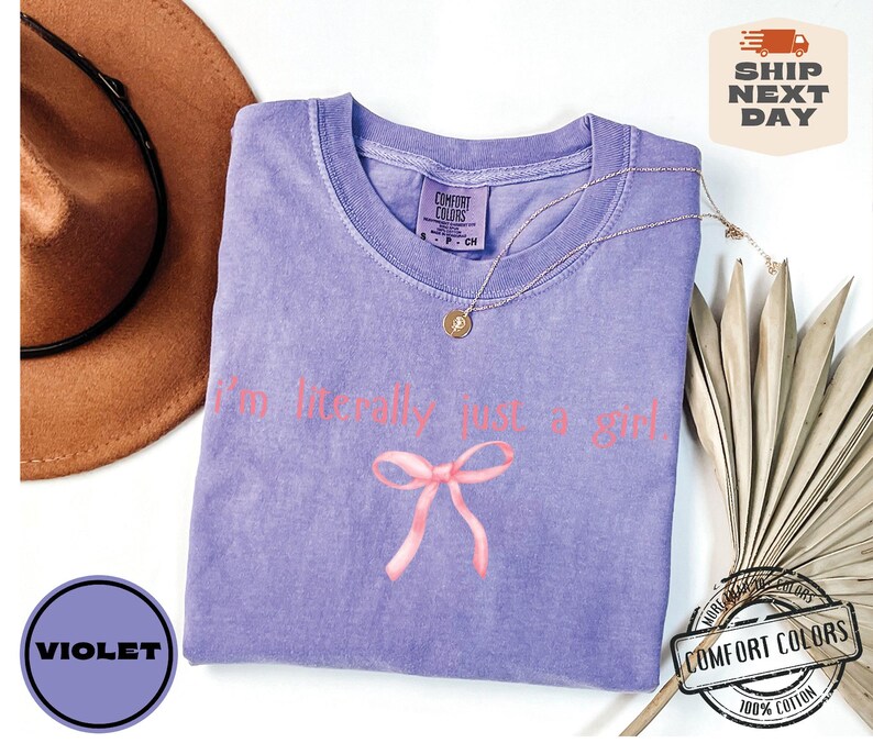 Puede incluir: Una camiseta morada con el texto "I'm literally just a girl!" en letras rosas y un lazo rosa. La camiseta est&aacute; hecha de 100% algod&oacute;n.