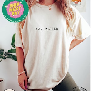 May include: A white t-shirt with the text "YOU MATTER" printed in black on the front. The shirt is being modeled by a person wearing it.