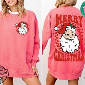 May include: A pink sweatshirt with a vintage-style Christmas graphic on the back. The graphic features a red and white Santa Claus with the text "Merry Christmas". The front of the sweatshirt has a smaller image of Santa Claus.