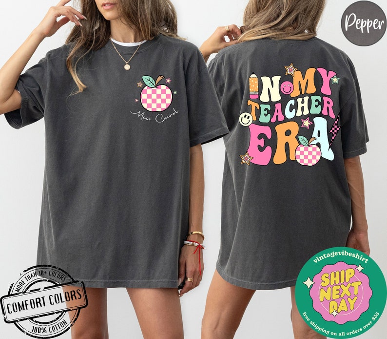 K&ouml;nnte beinhalten: Ein dunkelgraues T-Shirt mit einem pink-wei&szlig; karierten Apfel-Motiv und dem Text "Miss Carol" auf der Vorderseite. Die R&uuml;ckseite des Shirts zeigt eine bunte Grafik mit dem Text "In My Teacher Era".