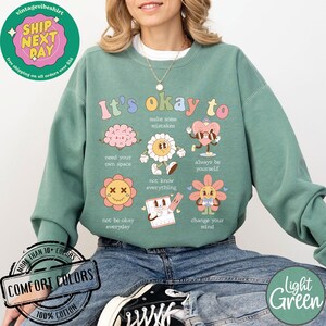 May include: Light green sweatshirt with a graphic print of colorful cartoon characters and the text "It's okay to" with phrases like "make some mistakes", "need your own space", "always be yourself", "not know everything", "not be okay everyday", and "change your mind".