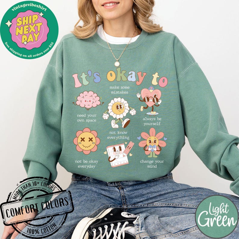 May include: Light green sweatshirt with a graphic print of colorful cartoon characters and the text "It's okay to" with phrases like "make some mistakes", "need your own space", "always be yourself", "not know everything", "not be okay everyday", and "change your mind".