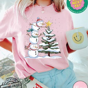 以下が含まれることがあります： 雪だるまの家族がクリスマスツリーを飾っているピンクのTシャツ。雪だるまの家族は、一番小さい雪だるまが下になるように積み重ねられています。木は緑色で、枝には白い雪が積もっています。背景は薄いピンク色で、白い雪の結晶が散りばめられています。