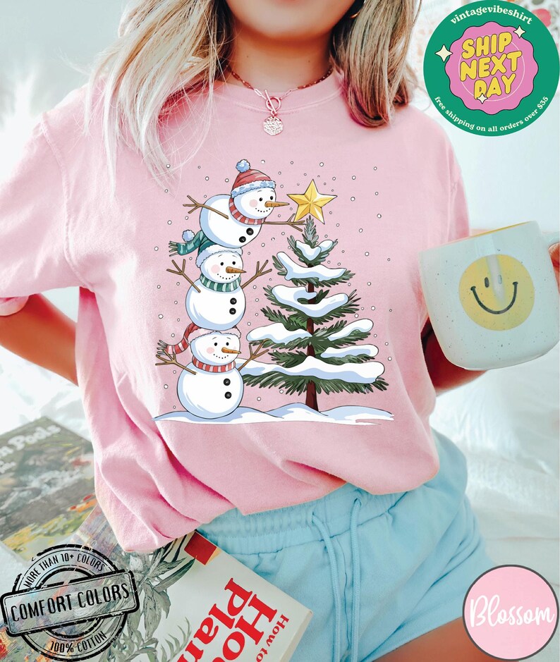 以下が含まれることがあります： 雪だるまの家族がクリスマスツリーを飾っているピンクのTシャツ。雪だるまの家族は、一番小さい雪だるまが下になるように積み重ねられています。木は緑色で、枝には白い雪が積もっています。背景は薄いピンク色で、白い雪の結晶が散りばめられています。