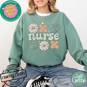 May include: Light green sweatshirt featuring the word "nurse" in a retro font, surrounded by flowers, hearts, and plasters. The sweatshirt is made from 100% cotton and is available in a range of colours.