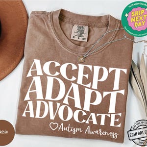 May include: Espresso brown t-shirt with the text "ACCEPT ADAPT ADVOCATE" in white. A small heart with the text "Autism Awareness" is below the text. The shirt is made of 100% cotton.