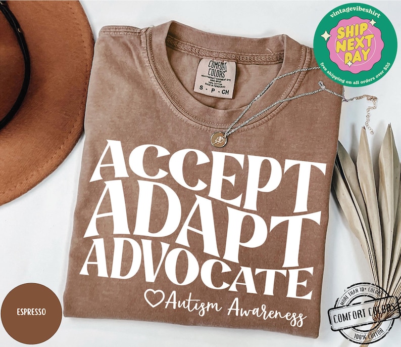 May include: Espresso brown t-shirt with the text "ACCEPT ADAPT ADVOCATE" in white. A small heart with the text "Autism Awareness" is below the text. The shirt is made of 100% cotton.