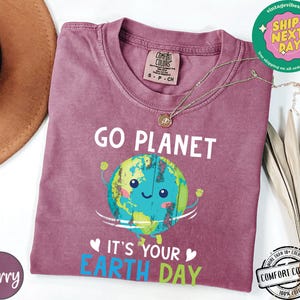 May include: A maroon t-shirt with a graphic of a smiling cartoon Earth holding up its arms. The text "Go Planet It's Your Earth Day" is printed on the shirt.