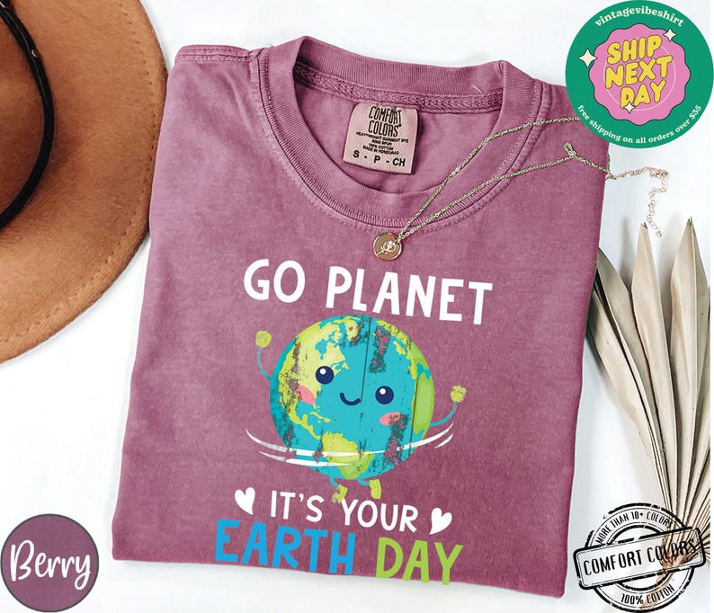 May include: A maroon t-shirt with a graphic of a smiling cartoon Earth holding up its arms. The text "Go Planet It's Your Earth Day" is printed on the shirt.