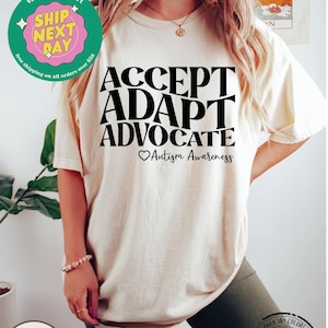 May include: A white t-shirt with black text that reads "ACCEPT ADAPT ADVOCATE" with a heart and the words "Autism Awareness" below. The shirt is being worn by a person.