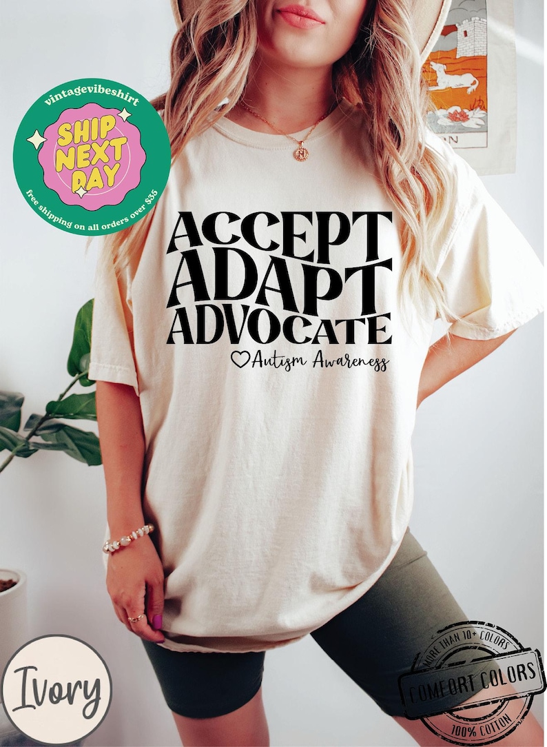 May include: A white t-shirt with black text that reads "ACCEPT ADAPT ADVOCATE" with a heart and the words "Autism Awareness" below. The shirt is being worn by a person.