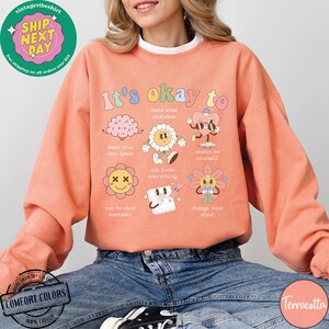 May include: Terracotta colored sweatshirt with a graphic print of cartoon characters and the text "It's okay to" with phrases like "make some mistakes", "need your own space", "not know everything", "always be yourself", "not be okay everyday", and "change your mind".