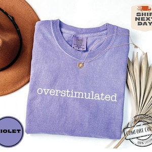May include: A violet Comfort Colors t-shirt with the word "overstimulated" in white. A gold necklace and a brown hat are next to the shirt. The shirt has a small tag that says "Comfort Colors".