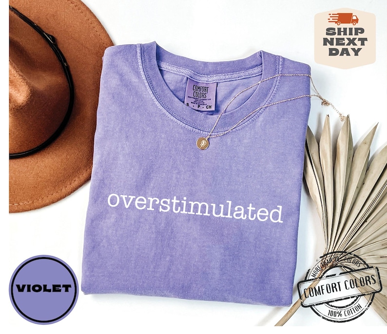 May include: A violet Comfort Colors t-shirt with the word "overstimulated" in white. A gold necklace and a brown hat are next to the shirt. The shirt has a small tag that says "Comfort Colors".