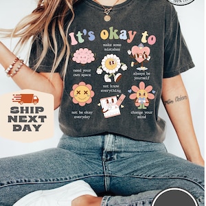 May include: A dark gray t-shirt with the text "It's okay to" and several cartoon illustrations. The illustrations include a brain, a flower, a heart, and a pencil. The text below each illustration includes phrases like "make some mistakes", "need your own space", "not know everything", "always be yourself", "not be okay everyday", and "change your mind".