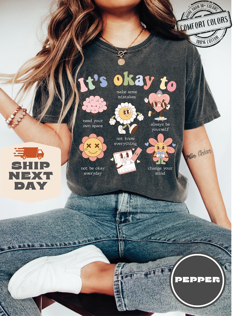 May include: A dark gray t-shirt with the text "It's okay to" and several cartoon illustrations. The illustrations include a brain, a flower, a heart, and a pencil. The text below each illustration includes phrases like "make some mistakes", "need your own space", "not know everything", "always be yourself", "not be okay everyday", and "change your mind".