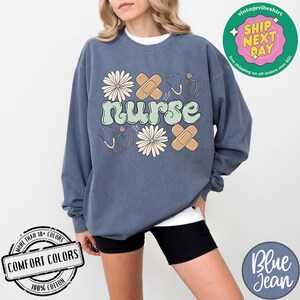 May include: A blue sweatshirt featuring the word "nurse" in green lettering, along with a design of flowers, bandages, and a stethoscope. The sweatshirt is made from 100% cotton and comes in a range of colours.