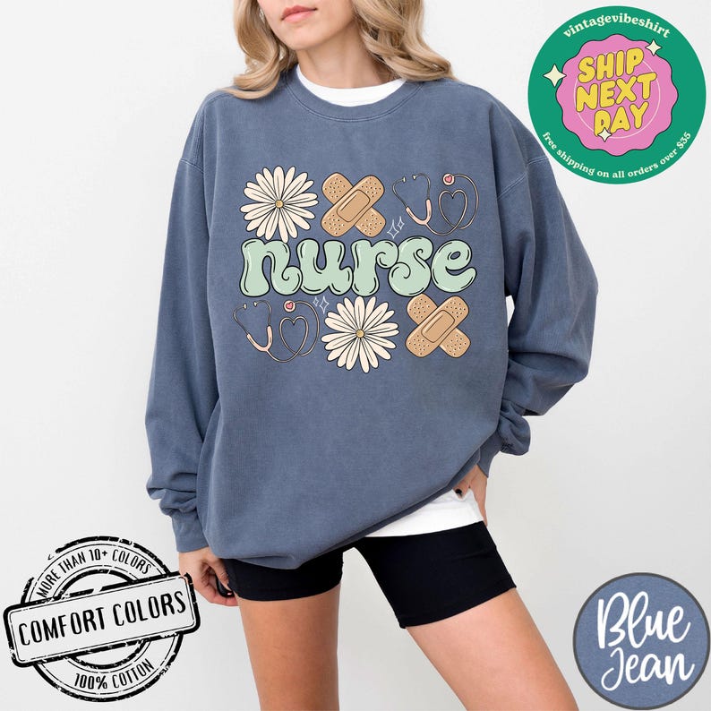 May include: A blue sweatshirt featuring the word "nurse" in green lettering, along with a design of flowers, bandages, and a stethoscope. The sweatshirt is made from 100% cotton and comes in a range of colours.