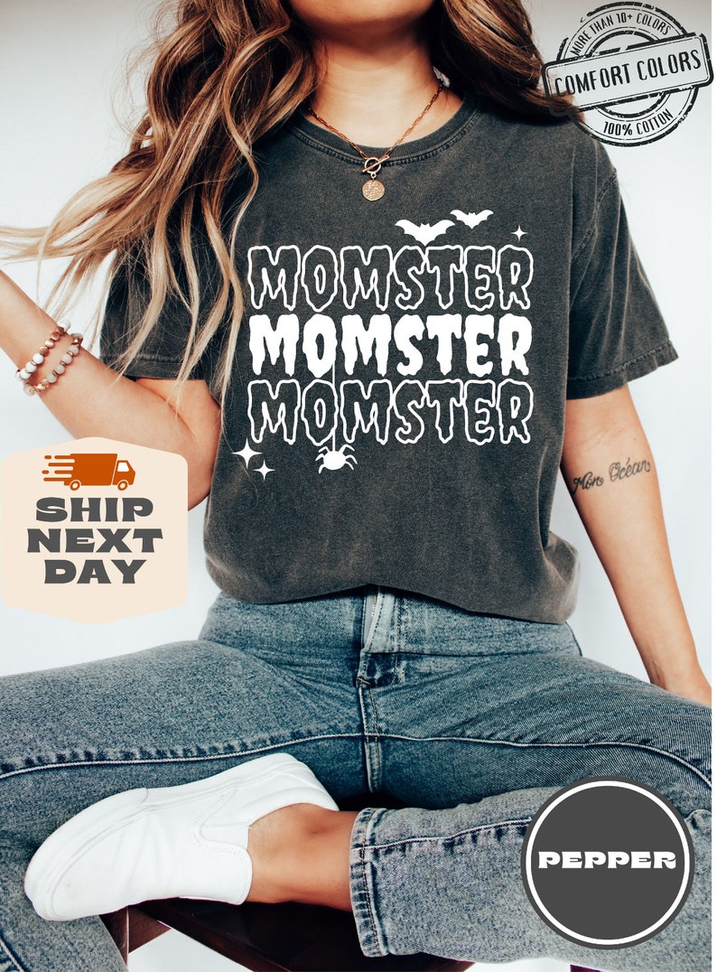 May include: A dark gray t-shirt with the text "Momster" repeated three times in white. The text is in a dripping font style. There is a small spider and a bat graphic above the text.