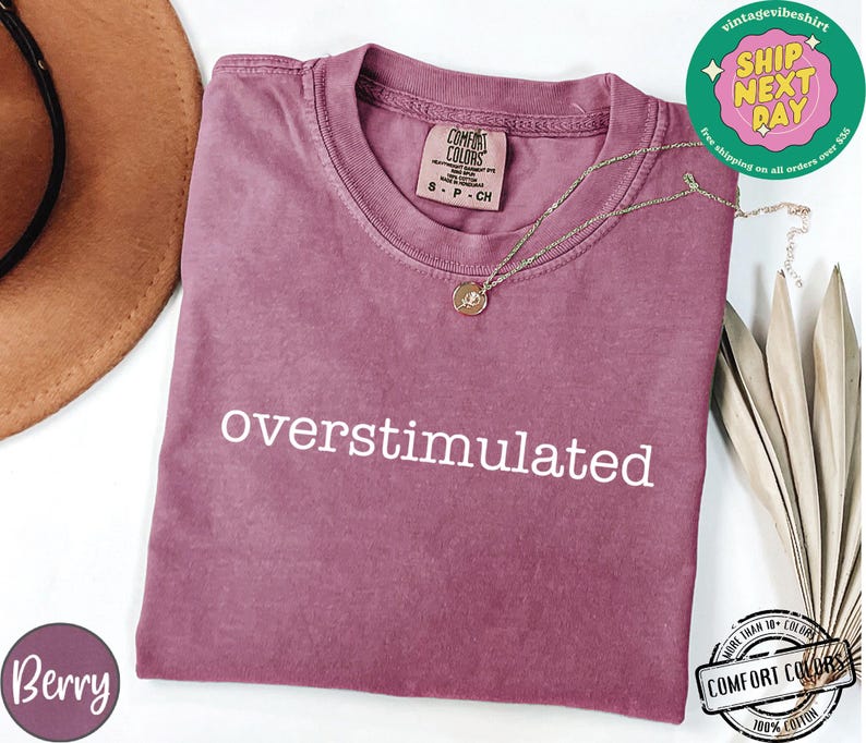 May include: Berry-colored t-shirt with the word "overstimulated" in white. The shirt has a crew neck and short sleeves. A gold necklace with a pendant is draped on the shirt. The shirt is made of Comfort Colors 100% cotton.