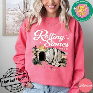 May include: A pink sweatshirt with a distressed white graphic that says "Rolling Stones Since AD 33". The graphic features a stone tomb with a cross and a white cloth draped over it.